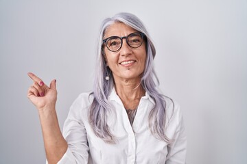 Wall Mural - Middle age woman with tattoos wearing glasses standing over white background with a big smile on face, pointing with hand and finger to the side looking at the camera.