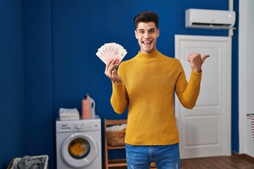 Wall Mural - Young hispanic man at laundry room holding shekels pointing thumb up to the side smiling happy with open mouth