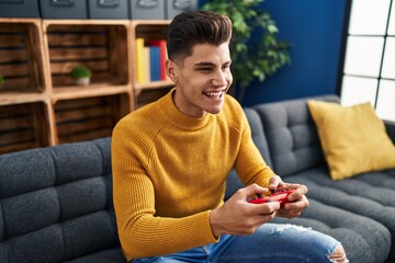 Wall Mural - Young hispanic man playing video game sitting on sofa at home