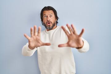 Poster - Handsome middle age man wearing casual sweater over blue background afraid and terrified with fear expression stop gesture with hands, shouting in shock. panic concept.