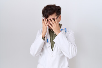 Sticker - Young non binary man wearing doctor uniform and stethoscope with sad expression covering face with hands while crying. depression concept.