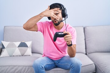 Poster - Hispanic young man playing video game holding controller sitting on the sofa peeking in shock covering face and eyes with hand, looking through fingers with embarrassed expression.