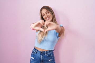 Poster - Young blonde woman standing over pink background smiling in love doing heart symbol shape with hands. romantic concept.