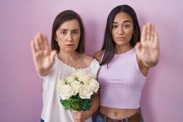 Wall Mural - Hispanic mother and daughter holding bouquet of white flowers doing stop sing with palm of the hand. warning expression with negative and serious gesture on the face.
