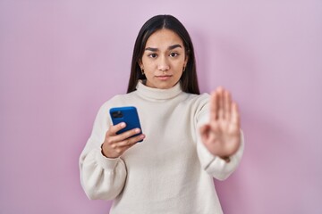 Wall Mural - Young south asian woman using smartphone with open hand doing stop sign with serious and confident expression, defense gesture