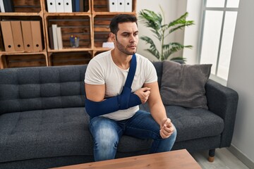 Sticker - Young hispanic man with beard wearing arm on sling sitting at therapy consult thinking attitude and sober expression looking self confident