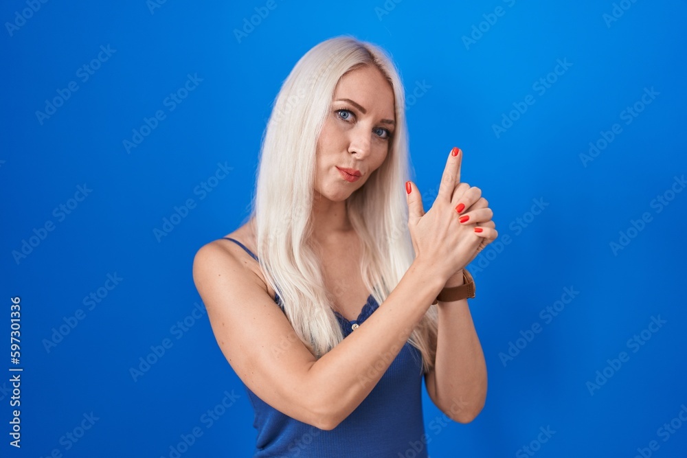 Caucasian woman standing over blue background holding symbolic gun with hand gesture, playing killing shooting weapons, angry face