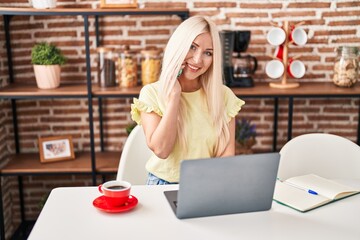 Wall Mural - Young blonde woman using laptop and talking on the smartphone sitting on table at home