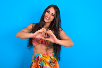 Wall Mural - beautiful brunette woman wearing swimwear over blue background smiling in love doing heart symbol shape with hands. Romantic concept.