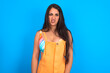 Portrait of dissatisfied brunette woman wearing orange overalls over blue studio background smirks face, purses lips and looks with annoyance at camera, discontent hearing something unpleasant