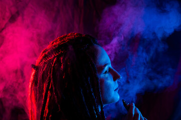 Profile of caucasian girl with dreadlocks smokes a vape in red blue light. 