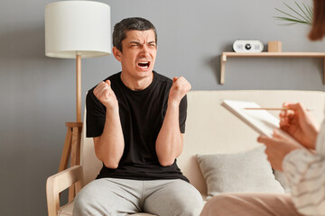 Angry aggressive young man in black T-shirt sitting on sofa at psychologist office, sharing feelings, thoughts with therapist woman, screaming with hate during therapy session.