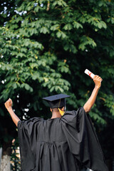 Afro american graduate from university in black mantle and hat stand with back outdoors among trees and holding higher education diploma with raised arms. Start in life, graduation, freedom.