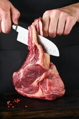 Wall Mural - Chef cutting steak beef. Mans hands hold raw steak Tomahawk on rustic wooden cutting board on black background. Cooking, recipes and eating concept. Selective focus.