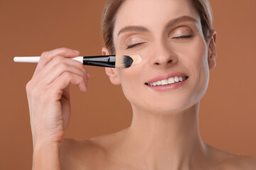 Wall Mural - Woman applying foundation on face with brush against brown background