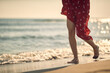 Shot of legs of young woman in red summer dress walking on beach by waves of water. Fashion, summertime, lifestyle concept.