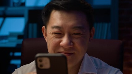 Happy Asian middle-aged man Korean adult businessman at night evening home office using mobile phone smiling with smartphone laughing browsing cellphone internet service online telephone app smile