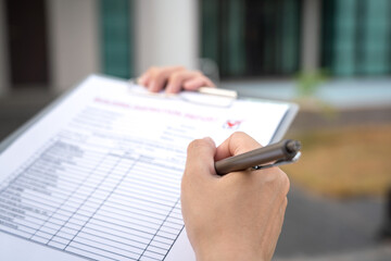 Wall Mural - Foreman's hand is using a pen checking on building inspection report form to QC building quality of the house (background). Construction industrial working scene concept. Close-up and selective focus.