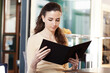 © amedeoemaja - Beautiful young woman at the coffee bar, ice cream shop or pastry shop holding a black menu on her hands. Portrait of brunette smiling girl looking at the menu of the coffee bar.