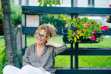 Portrait of young mature woman having relax and enjoying home outdoor garden sitting and smiling. One female people serene relaxing outside home. Happy adult lady wearing eyeglasses sit down. Cheerful