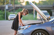 © bilanol - Helpless woman standing near her car with open bonnet inspecting broken motor. Young female driver having trouble with vehicle