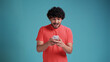 young latin man looks delighted into smartphone and says wow. Dressed in casual on a blue studio background