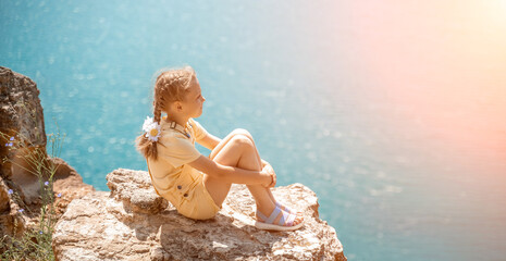 Happy girl perched atop a high rock above the sea, wearing a yellow jumpsuit and braided hair, signifying the concept of summer vacation at the beach.