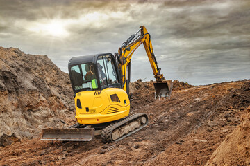 Wall Mural - Mini excavator at the construction site on the edge of a pit against a cloudy blue sky. Compact construction equipment for earthworks. An indispensable assistant for earthworks.