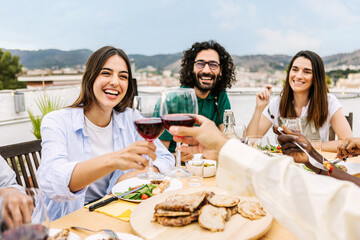 Wall Mural - Happy diverse group of friends enjoying dinner barbecue in summer at home terrace. Two young people cheering with red wine at bbq party. Food and family moments concept