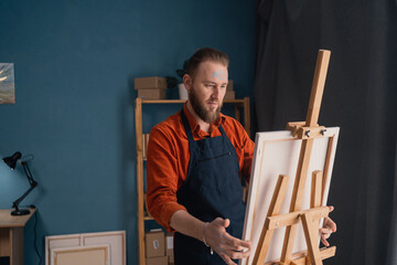 portrait caucasian bearded male artist in an apron in a home studio stands near an easel holds a canvas in his hands and looks at the picture contemptuously. hobby and vacation concept