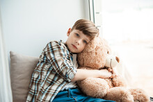 Child Boy Hugging Teddy Bear Sitting Alone At Window Behind Transparent Day Curtains And Looking Out From Home. Waiting Concept. Close Up