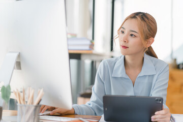 A focused and professional millennial Asian businesswoman in casual attire is analyzing financial data on her desktop computer and tablet at her desk in a modern office.