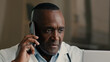 © Yuliia - African American doctor adult man work with computer at hospital use modern medical technology call patient remote by mobile phone male professional therapist consulting distant telemedicine concept