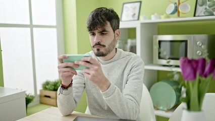 Sticker - Young hispanic man playing video games with smartphone at dinning room