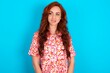 © Jihan - Caucasian redhead woman wearing floral dress over blue background with thoughtful expression, looks away keeps hands down bitting his lip thinks about something pleasant.