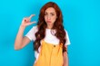 Young redhead woman wearing orange overall over blue background purses lip and gestures with hand, shows something very little.