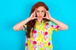 Serious concentrated Young redhead woman wearing colorful shirt over blue background keeps fingers on temples, tries to ease tension, gather with thoughts and remember important information for exam