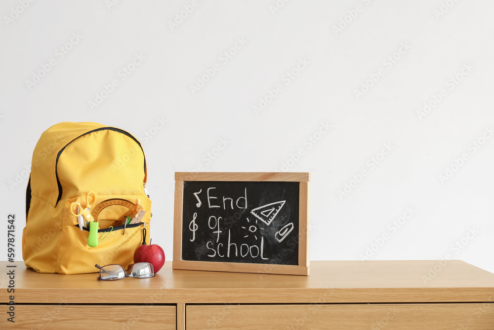 Chalkboard with text END OF SCHOOL, apple and backpack on table near light wall