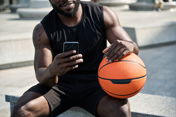 Fit sporty young African black ethnic man sitting outdoors holding basketball ball and mobile phone using apps, looking at smartphone, resting after street sport game with cellphone. Closeup