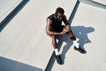 Fit Sporty Young Black Man Sitting On Concrete Urban Stairs Holding Phone Using Mobile Apps Listening Music. Strong African Ethnic Guy Wearing Headphones Looking At Smartphone Outdoors. Top View