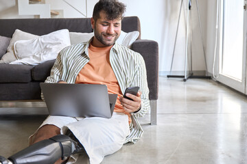 Happy young man with disability leg prosthesis sitting on floor at home holding laptop and mobile phone working, browsing web, searching online. People with amputation disabilities everyday life.