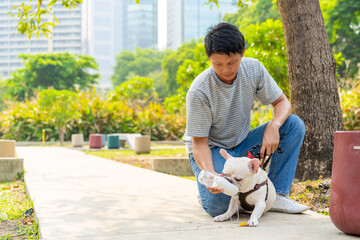 Asian man giving water to french bulldog breed during walking together at pets friendly dog park. Domestic dog with owner enjoy urban outdoor lifestyle on summer vacation. Pet Humanization concept.