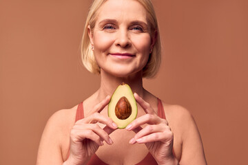 Wall Mural - A beautiful smiling senior woman holds half an avocado with two hands on a beige background. Beauty, health care.