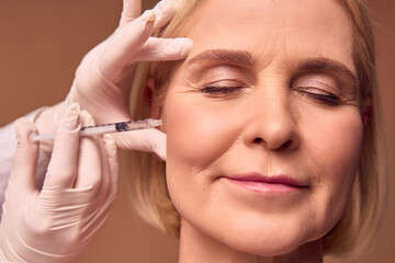 Close-up portrait of an adult woman with closed eyes on a beige background. Hands in white gloves and a medical gown make an injection in the cheekbone. Smoothing out wrinkles. Aesthetic medicine.