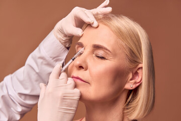 Portrait of an older adult woman with closed eyes doing a beauty injection between the eyebrows. Hands in white gloves and a medical coat hold a syringe.Smoothing out wrinkles.