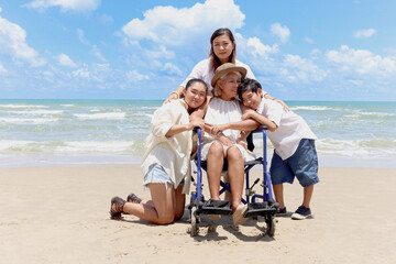 Happy disabled senior elderly woman in wheelchair hugging with her family on tropical beach. Asian grandma, daughter and grandchild boy resting and relaxing together on summer holiday vacation.