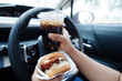 © manassanant - Asian woman driver hold and eat hamburger in car, dangerous and risk an accident.