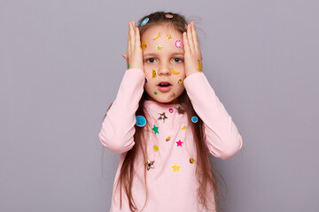 Shocked scared little girl covered colorful kid's stickers standing isolated over gray background keeps hand on head sees something frightened looking with big full of scary eyes.