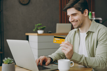 Wall Mural - Young smiling happy fun man wears casual clothes sits alone at table in coffee shop cafe indoors work using laptop pc computer hold credit bank card shopping online. Freelance mobile office concept.