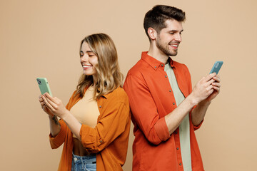 Wall Mural - Side view young couple two friends family man woman wear casual clothes hold in hand use mobile cell phone together stand back to back isolated on pastel plain light beige background studio portrait.